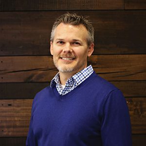 A smiling individual wearing a blue sweater and a checkered shirt stands against a wooden backdrop.
