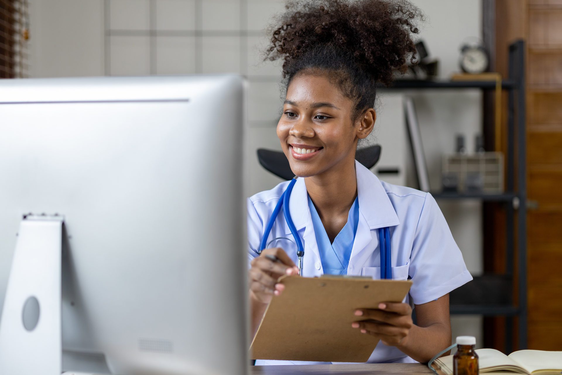 A smiling healthcare professional in scrubs is holding a clipboard and looking at a computer screen.