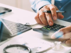 A person in a light blue shirt is using a calculator and writing notes, surrounded by financial documents and a laptop.