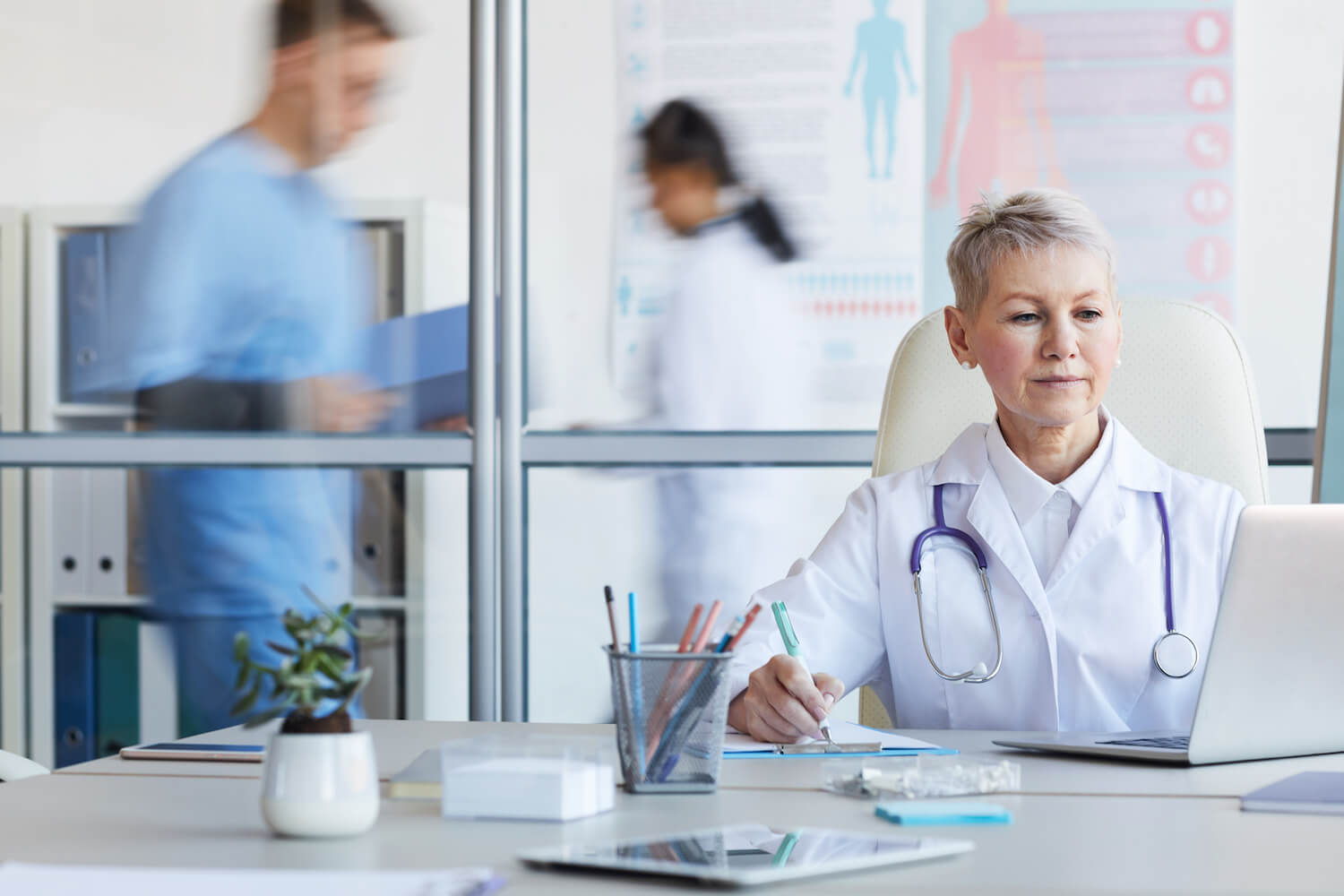 A healthcare professional sits at a desk with medical equipment, reviewing paperwork, while colleagues engage in activities in the background.